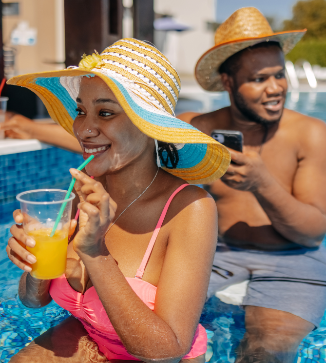Woman in a sunhat sipping a drink at a pool, man with phone behind her.