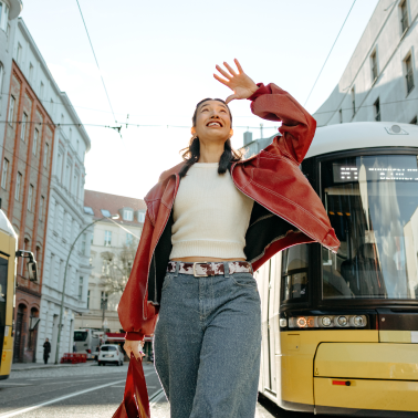 Woman smiling and shielding her eyes while walking past a yellow tram on a city street.