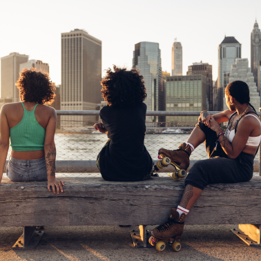 Three friends with roller skates sitting on a bench, looking at a city skyline.