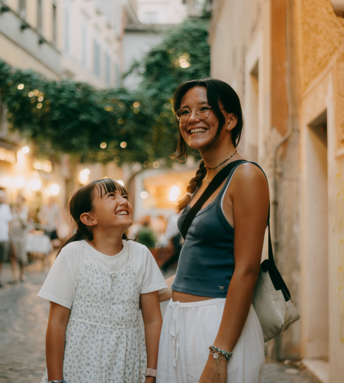 Two smiling girls standing in a cobblestone street with hanging greenery.