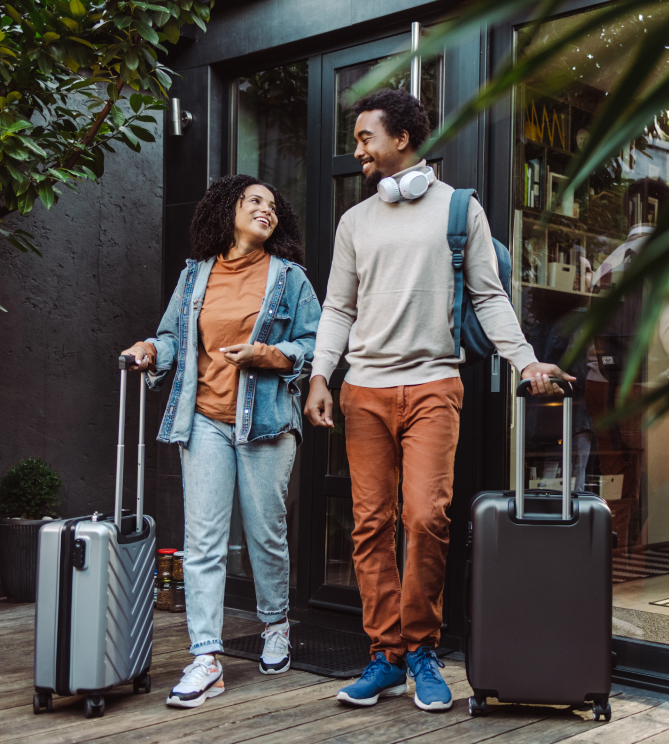 Couple smiling and walking out of a hotel with rolling suitcases.