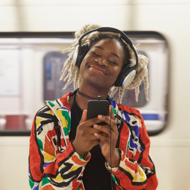 Person wearing headphones, smiling while using a phone on a subway platform.