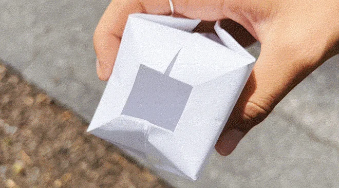 Hand squeezing a folded origami water balloon made from white paper.