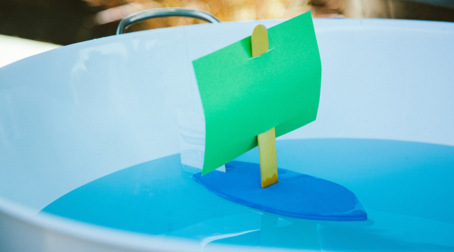 A small paper sailboat floating in a water-filled container with a green sail.