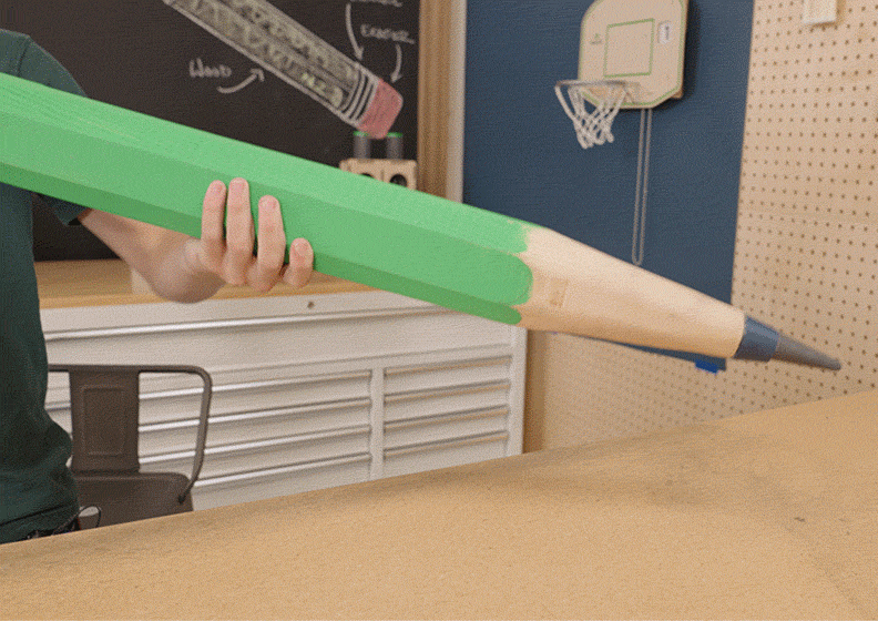 GIF: Giant Pencil in Action | (GIF shows a child lifting and presenting a giant green pencil in a classroom.)