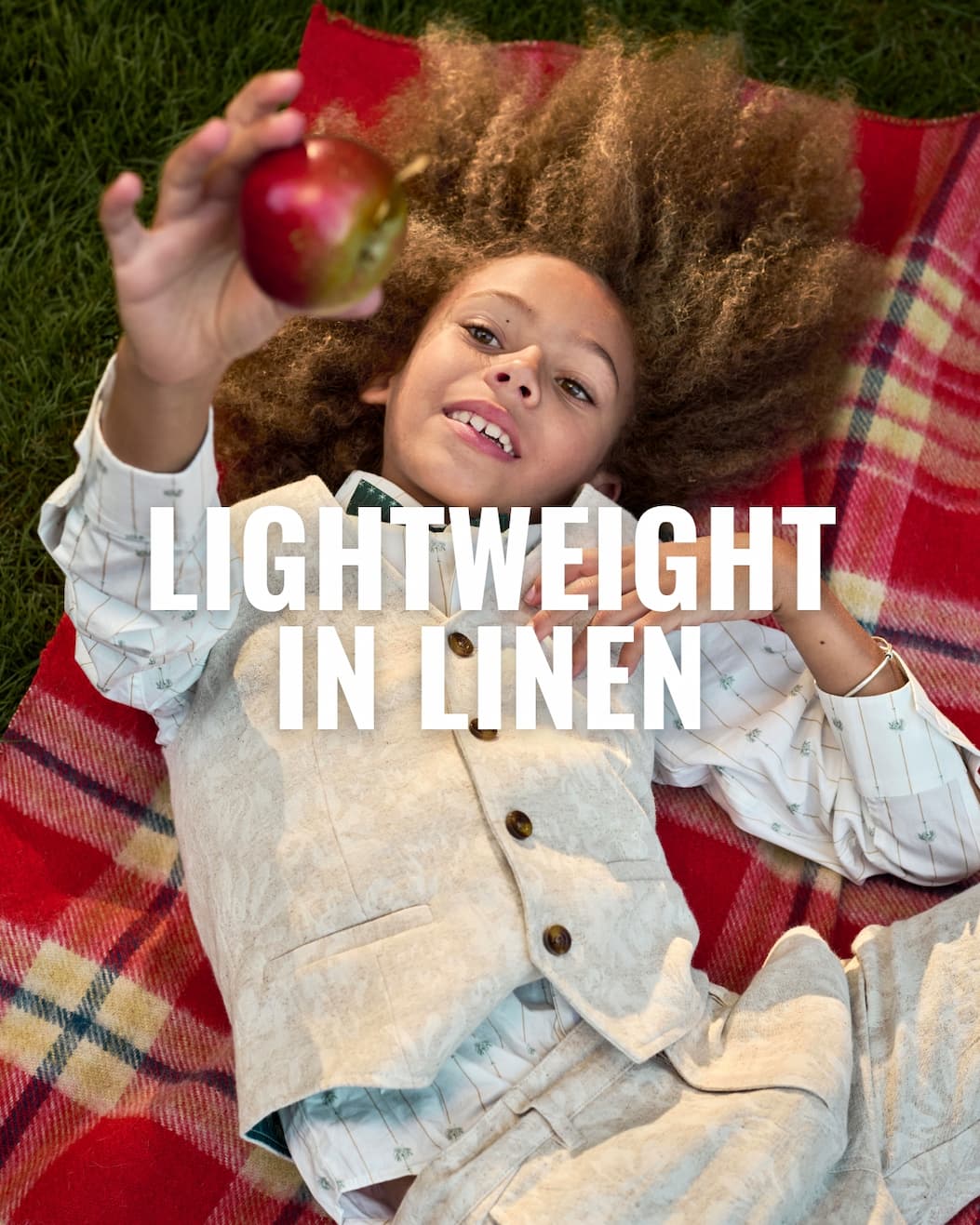 Boy in Beige Lightweight Linen Suit Laying on Red Blanket Holding a Red Apple