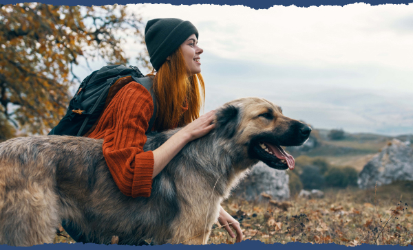 A hiker, wearing a beanie and a backpack, and her fluffy medium-sized dog are enjoying a picturesque view of rolling hills.