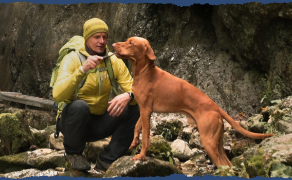 A hiker in a yellow jacket and beanie is feeding his excited dog a treat. They’re in a shaded area with moss-covered rocks.