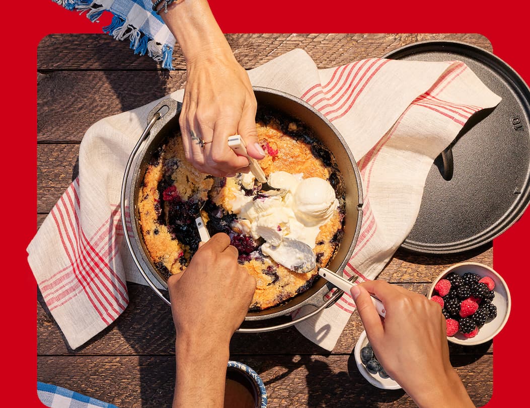 Image on red background, hands reaching over to share a berry dump cake in cast iron pan.