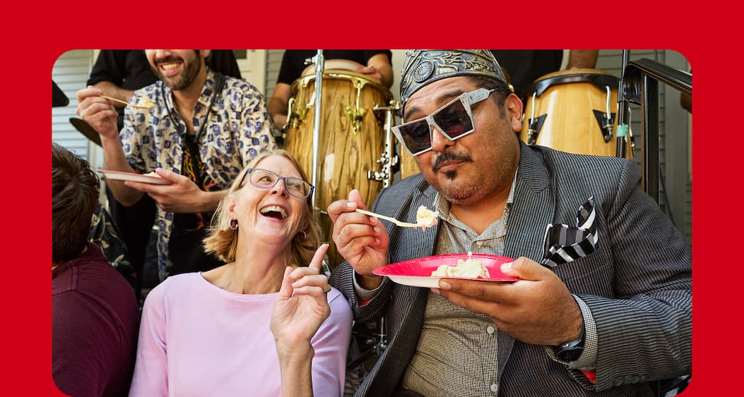 A woman and man sitting on a porch together happily eating cake while a live band plays in the background.