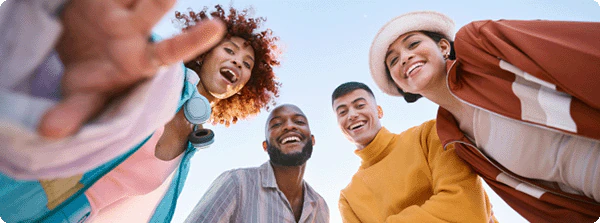 Low-angle shot of four smiling young adults of diverse backgrounds looking down at the viewer. They appear to be outdoors with a light blue sky in the background.