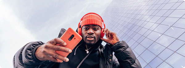 Low-angle photograph of a man wearing an orange knit cap and headphones, holding an orange phone up towards the camera. He is in front of a building with a glass facade.