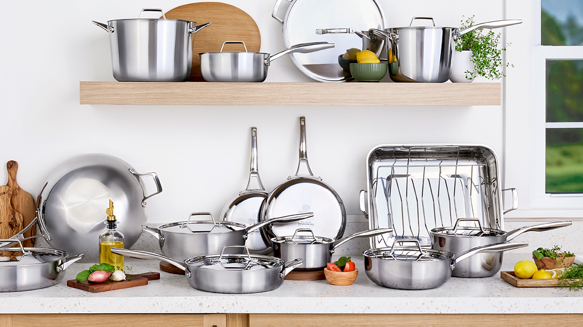 Stainless steel cookware set including pots, pans, and lids arranged on a kitchen counter with fresh ingredients and utensils nearby.