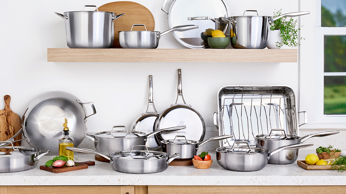 Stainless steel cookware set including pots, pans, and lids displayed on a kitchen countertop with fresh vegetables and herbs.
