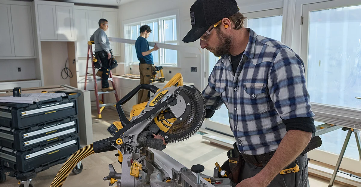 A man operates a DEWALT miter saw, model DCS714B, cutting a piece of wood inside a home under renovation. Two other workers are in the background installing trim, with tools and equipment visible in a bright kitchen setting.