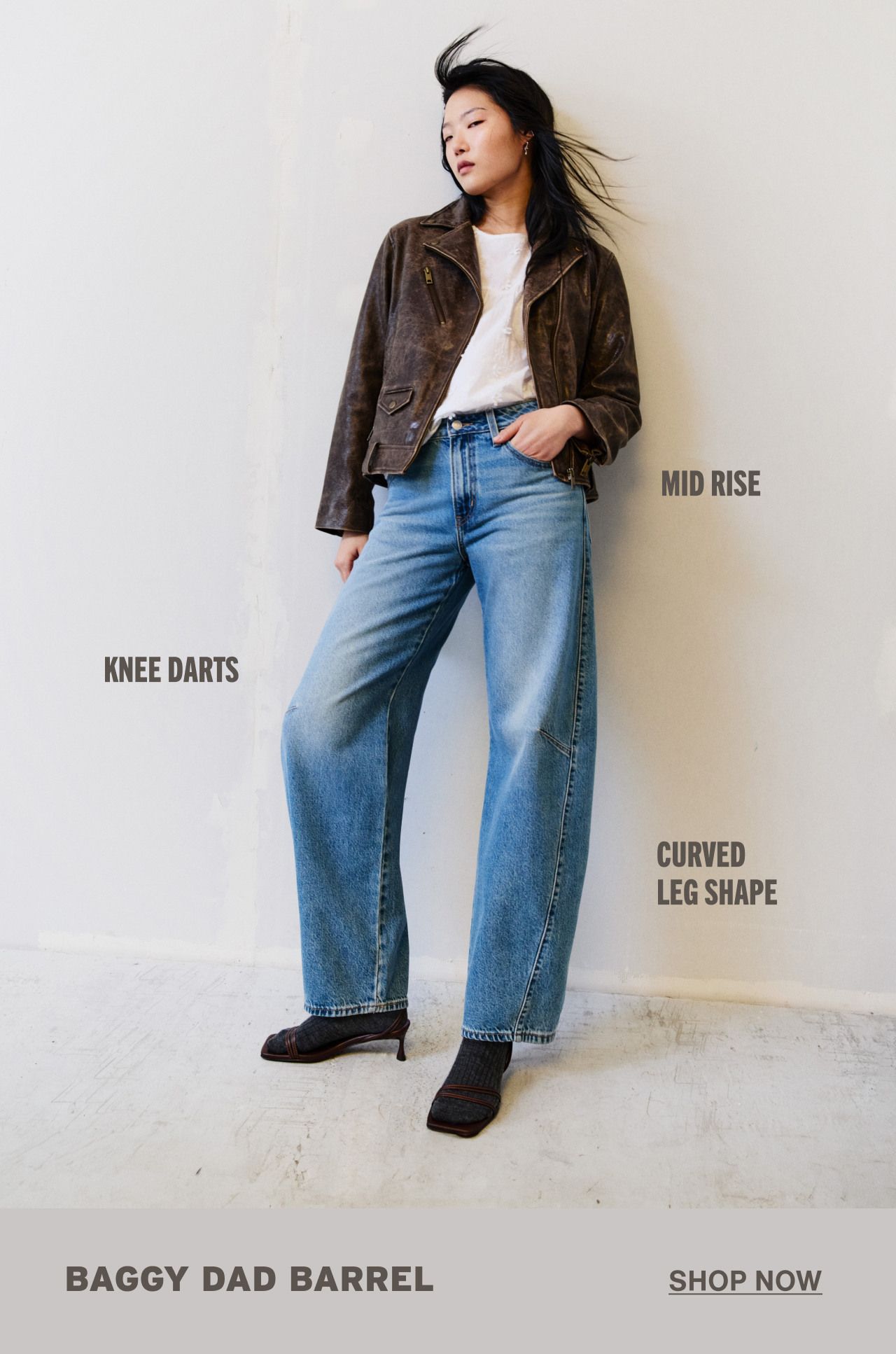 Person wearing a white shirt, brown jacket, and medium wash jeans leaning against the wall.