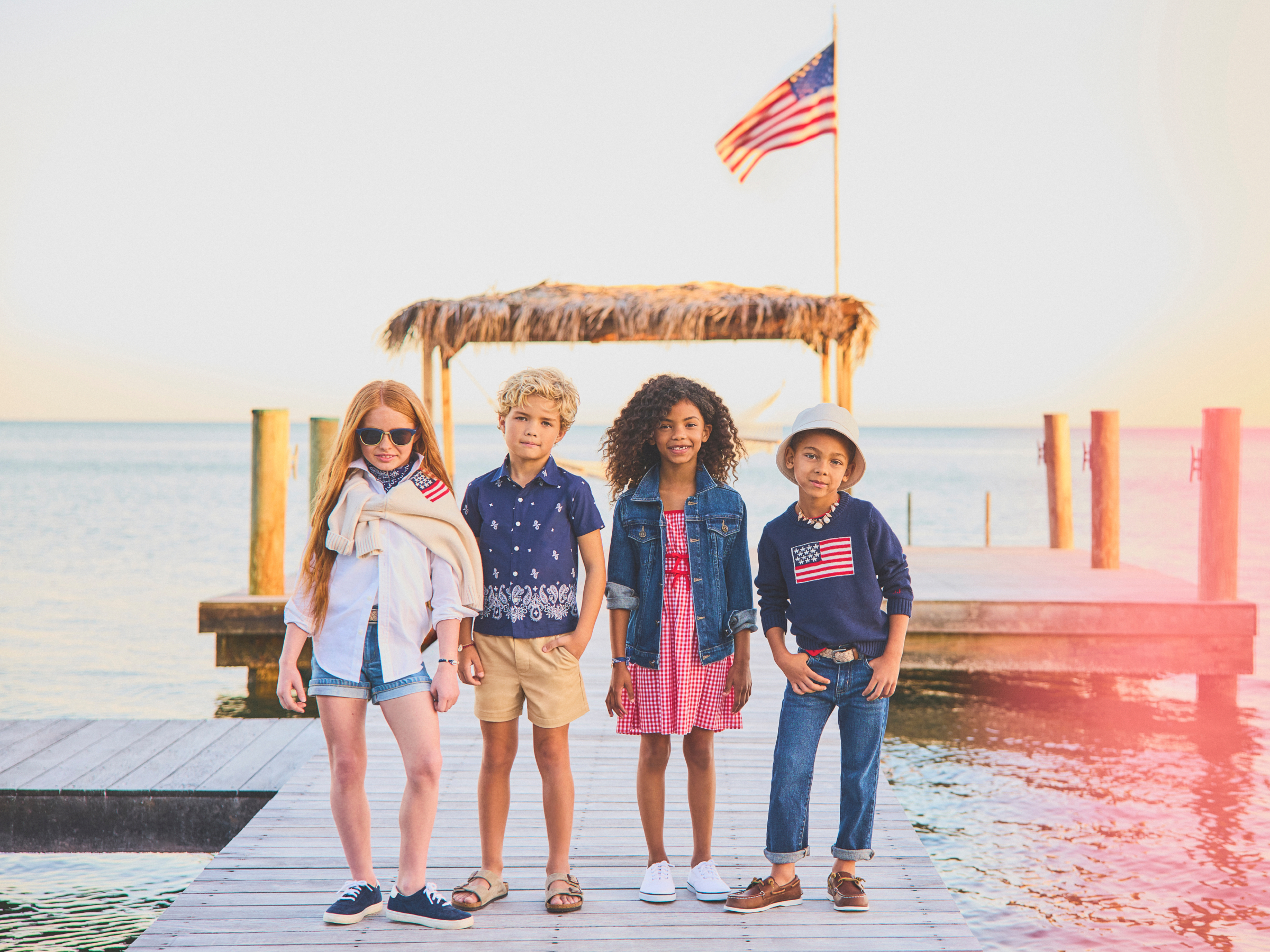 Four kids posing on a waterfront dock with an American flag in the background at sunset