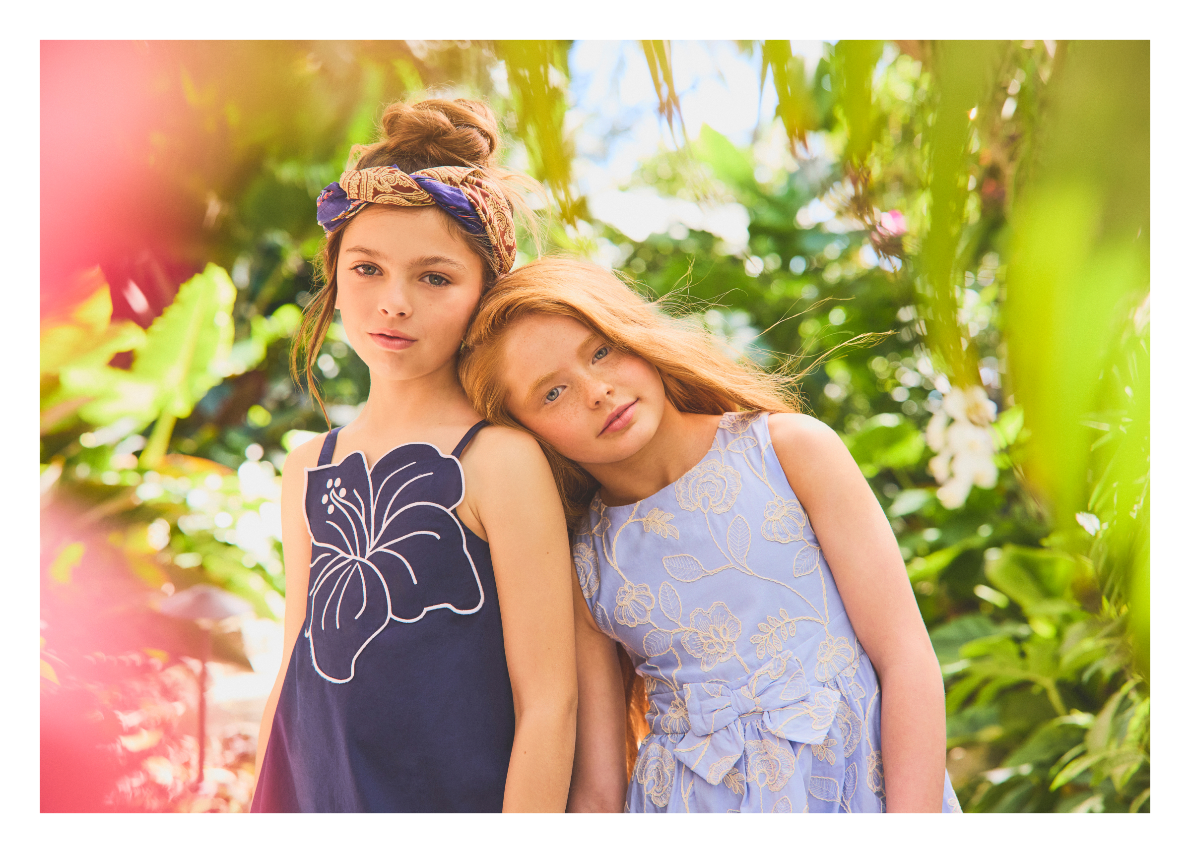 Two young girls in floral summer dresses posed among tropical greenery