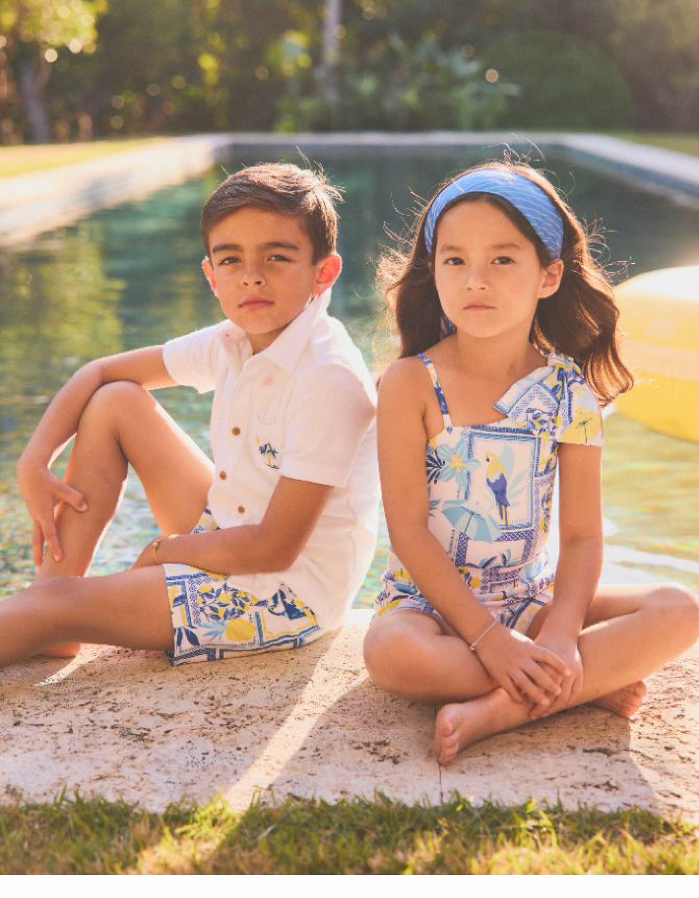 Two children sitting by a pool wearing coordinating blue, white, and yellow printed resort wear