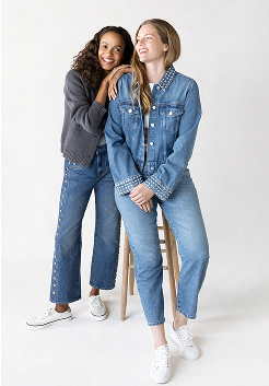 Two smiling women, one is sitting on a wooden stool and the other is leaning on her. The woman on the stool is wearing a studded denim jacket and straight-leg jeans paired with the Jump Kick Leather Lace-Ups in white. The other woman is wearing a grey cardigan and studded wide-leg denim paired with the Jump Kick T-Toe Leather/Suede Hair Calf Heel Lace-Ups in off white/beige.