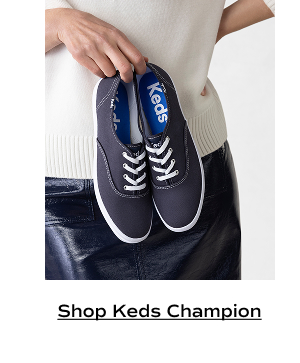 A woman in a cream sweater and navy leather skirt holding a pair of Keds Champion Canvas Lace-Ups in navy. Shop Keds Champion.