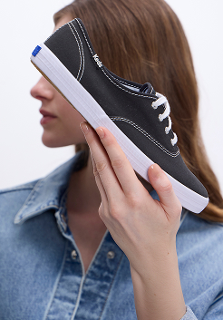 A woman in a denim button-up holding the Keds Champion Canvas Lace-Up in black.