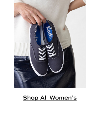 A woman in a cream sweater and navy leather skirt holding a pair of Keds Champion Canvas Lace-Ups in navy. Shop All Women's.