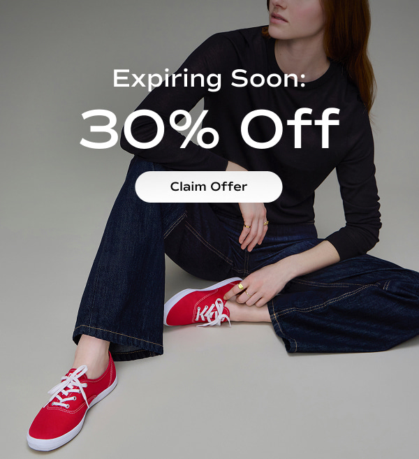 A woman sitting wearing a black long-sleeved T-shirt and dark-wash denim jeans paired with the Keds Champion Canvas Lace-Ups in red. Text over the image says 30% Off​ is expiring soon. Claim Offer​.​
