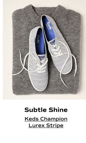 An overhead view of the Keds Champion Chambray Lurex Stripe in vintage indigo positioned on a grey wool sweater. The text says Subtle Shine. Shop the Keds Champion Lurex Stripe.