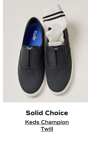 An overhead view of the Keds Champion Twill Round Toe Slip-On in charcoal with a ribbed white sock in one shoe. The text says Solid Choice. Shop the Keds Champion Twill.