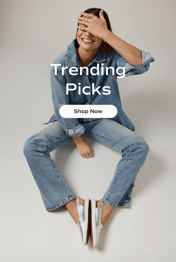 A woman smiling, sitting on the floor. She is wearing an all-denim outfit paired with the Point Slingback Washed Metallic Leather flats in silver. Text on the image says Trending Picks. Shop Now.