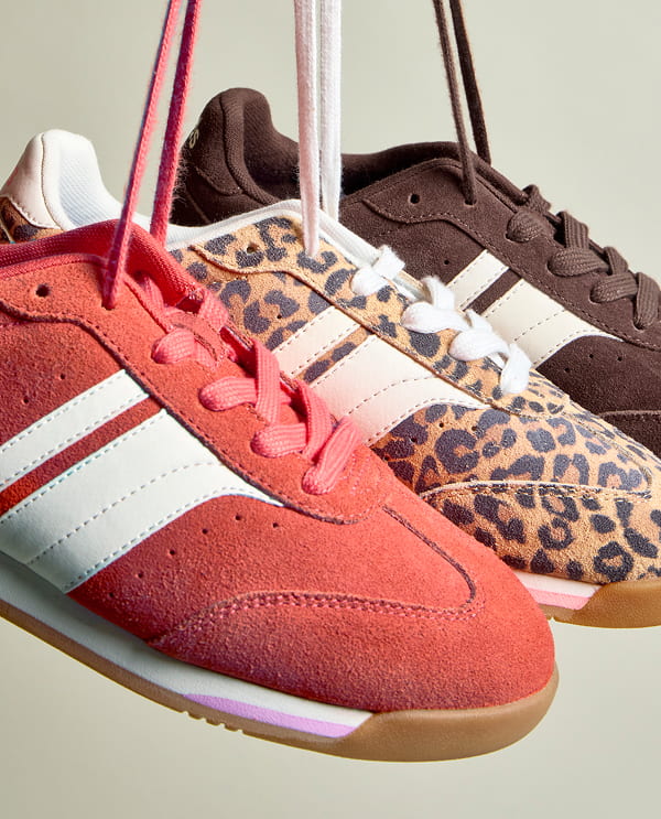 All three colors of the suede Revival—orange, tan leopard print, and chocolate—are shown. The three sneakers are being held up together by their laces, suspended in the air.