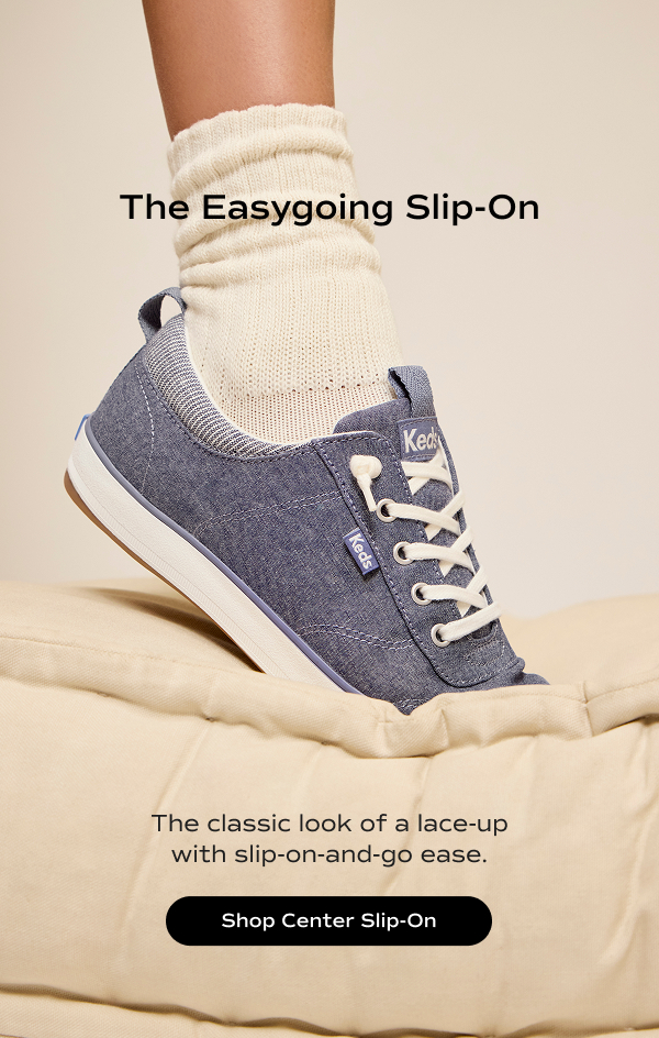 The Center III Chambray Slip-On in blue on foot paired with scrunched off-white sock stepping toe first into a plush comforter. The text says The Easygoing Slip-On. The classic look of a lace-up with slip-on-and-go ease. Shop Center Slip-On.