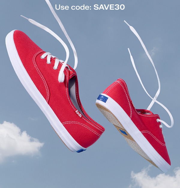 A pair of Keds Champion Canvas Lace-Ups in red floating in front of a background of a blue sky and puffy white clouds.
