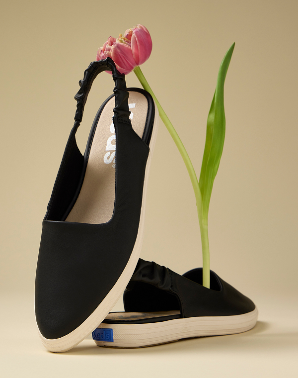 A pair of Point Slingback Leather flats in black shown on a beige background sitting with a long-stemmed pink tulip.