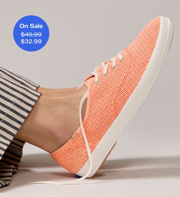 A closeup on a pair of Keds Champion Cotton Mesh Lace-Ups in neon coral shown on foot paired with black-and-beige pinstriped pants. A callout in the corner of the image notes that this sneaker was $49.99 and is now on sale for $32.99 for a limited time.