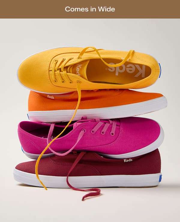 A brown banner with white text says Comes in Wide. A stack of four brightly colored Keds Champion Canvas Lace-Ups—dark red, raspberry, orange, and yellow—stacked on top of one another.