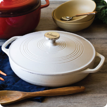 White enameled cast iron braiser with lid on a wooden table with bowls and utensils