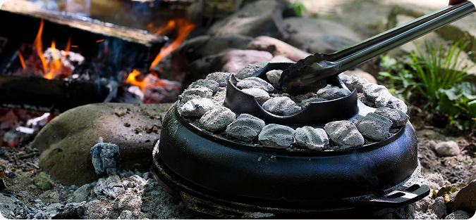 Cook-It-All being covered with hot charcoals next to a campfire