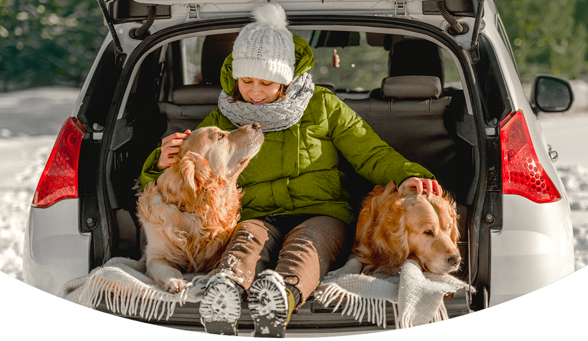 A woman bundled up on a snowy day sitting in her open back car with her two golden retrievers cozying up.