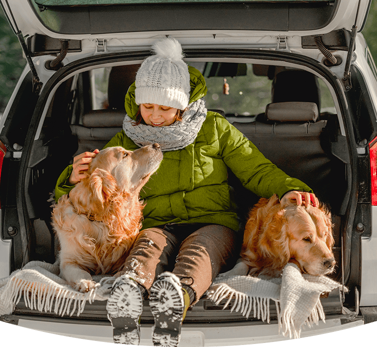 A woman bundled up on a snowy day sitting in her open back car with her two golden retrievers cozying up.