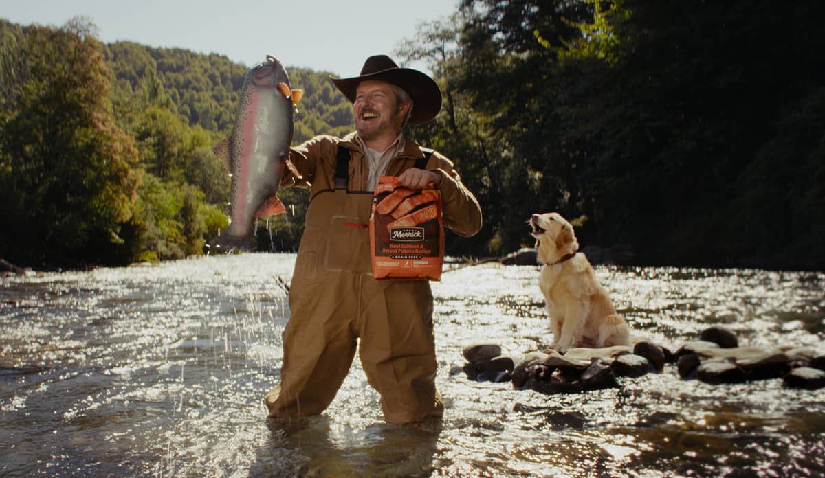 Cowboy in the river catching a fish in one hand while holding a bag of Merrick Real Salmon and Sweet Potato Recipe dry dog food in the other hand as a lab is barking in the back.