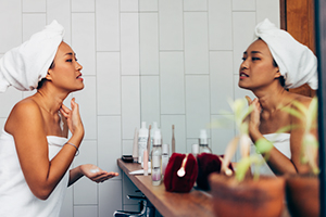 female presenting person looking in a mirror with towel on rubbing cream on her face and neck