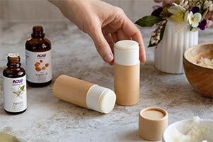 hand reaching down to pick up a tube of balm. Bottle of NOW Neroli Essential Oil and NOW Solutions Argan Oil 