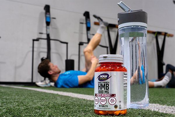 Bottle of NOW Sports HMB next to a bottle of water in front of a man working out in a gym 
