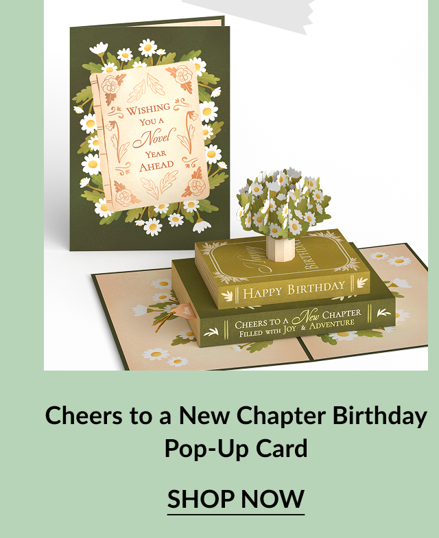 Birthday card styled like a stack of books with floral details and the message Wishing You a Novel Year Ahead on the cover. Inside reveals a pop-up tree growing from stacked books labeled Happy Birthday and Cheers to a New Chapter. Call-to-action below reads Cheers to a New Chapter Birthday Pop-Up Card – Shop Now.