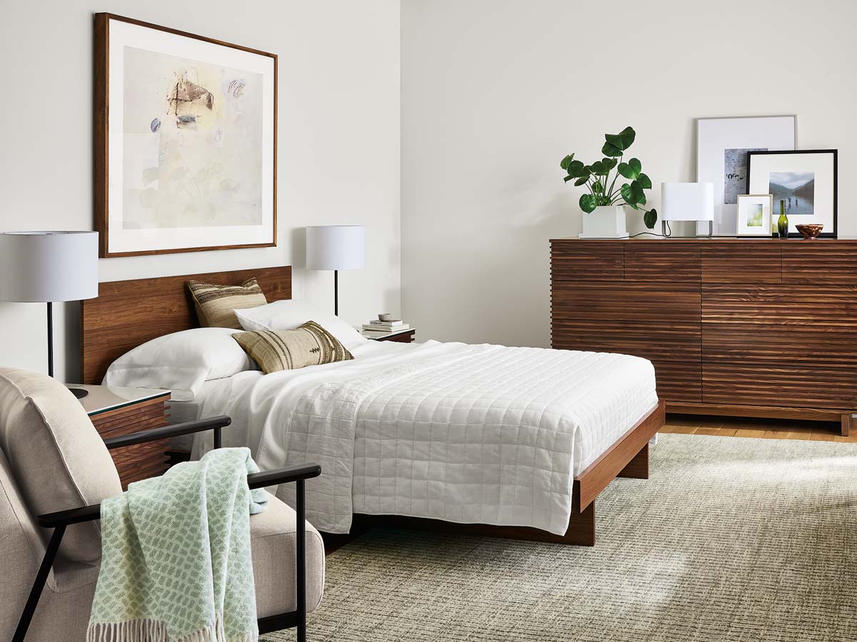 Anton bed with a wood headboard, white bedding, Coles dresser on the right, and nightstand with neutral-toned accent chair on the left.