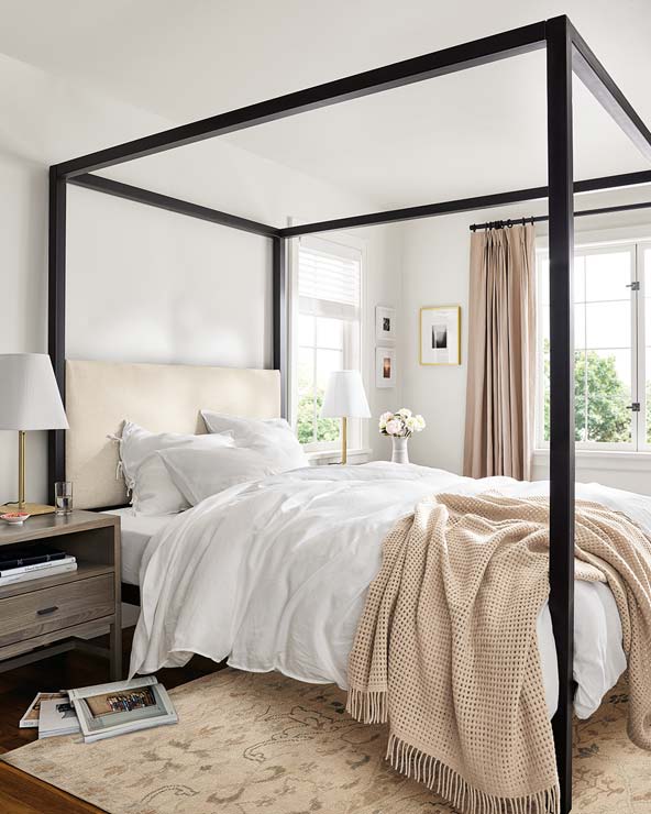 Canopy bed with an upholstered headboard, soft white bedding, beige knit throw, and nightstand with a modern lamp.