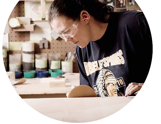 Artisan wearing safety glasses sanding smooth wood surface in workshop with organized tools in background.