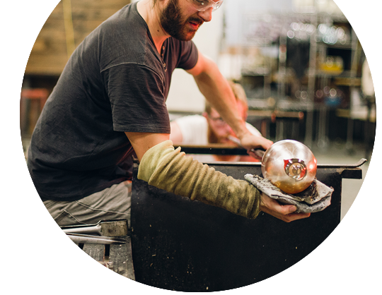Glassblower shaping molten glass on metal rod, forming round vessel under warm light in studio.